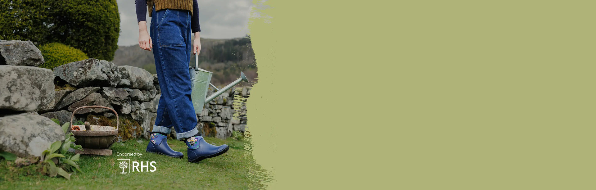 Woman holding a metal watering can wearing a pair of Muck Boots Muckster II Ankle Boots walking in front of a dry stone wall.