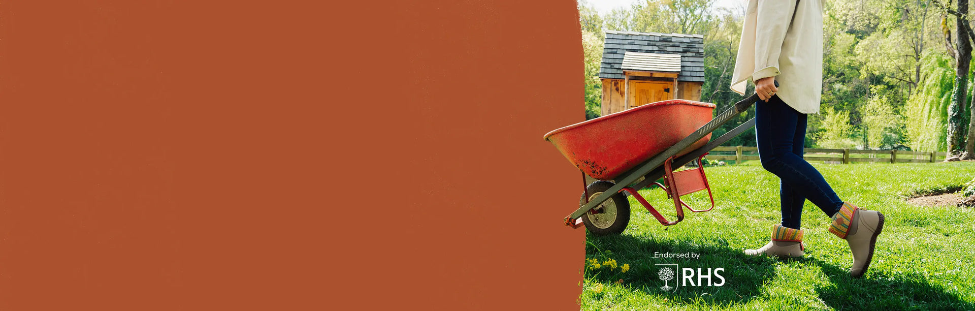 Person wearing a pair of Muck Boots Women's RHS Muckster II Short Boots pushing a red wheelbarrow with a small wooden shed in the background, featuring the RHS logo