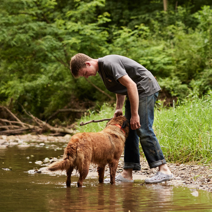 Boy wearing a pair of Muck Boots Muckster Clogs, playing with a dog in a stream. Text reads 'Muck Boot's Dog Walking Community'