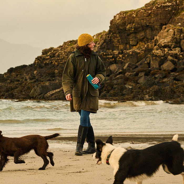 Woman walking along a beach with two dogs. She's wearing a beanie bat, long coat and a pair of Muck Boots Wellingtons