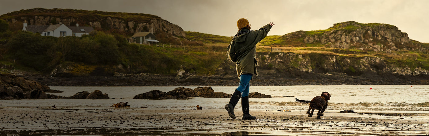 Woman wearing a pair of Muck Boots wellingtons on a beach, throwing a ball for a dog to chase