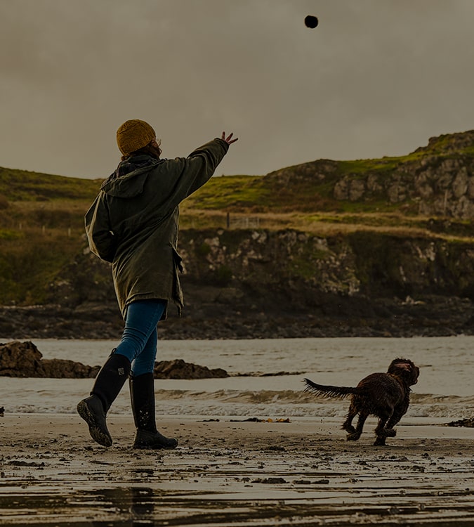 Woman wearing a pair of Muck Boots wellingtons on a beach, throwing a ball for a dog to chase