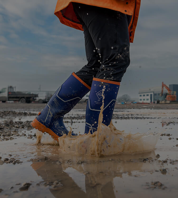 Person walking through a wet and muddy building site wearing a pair of blue Muck Boots Grit S5 Wellingtons