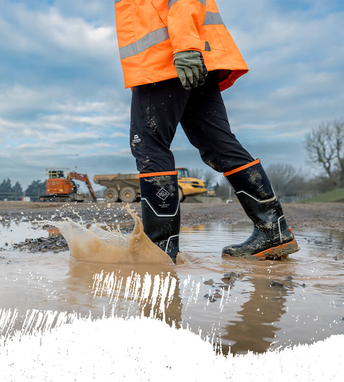 A man walking through muddy puddles on a building site, wearing a pair of Muck Boots Grit Safety Wellingtons