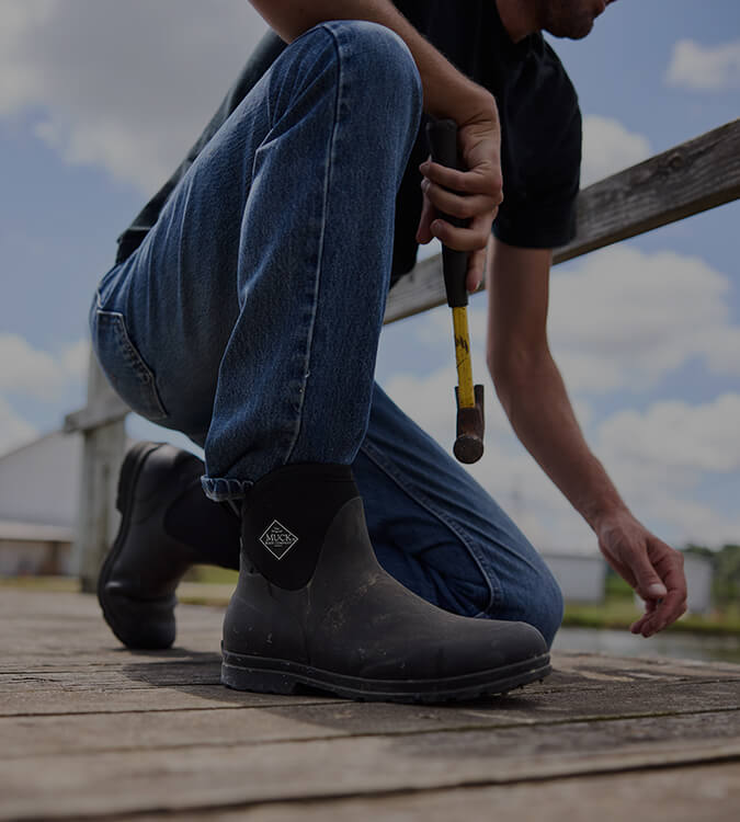 Man crouching down on one knee, holding a hammer in his right hand and wearing a pair of Muck Boots Originals.