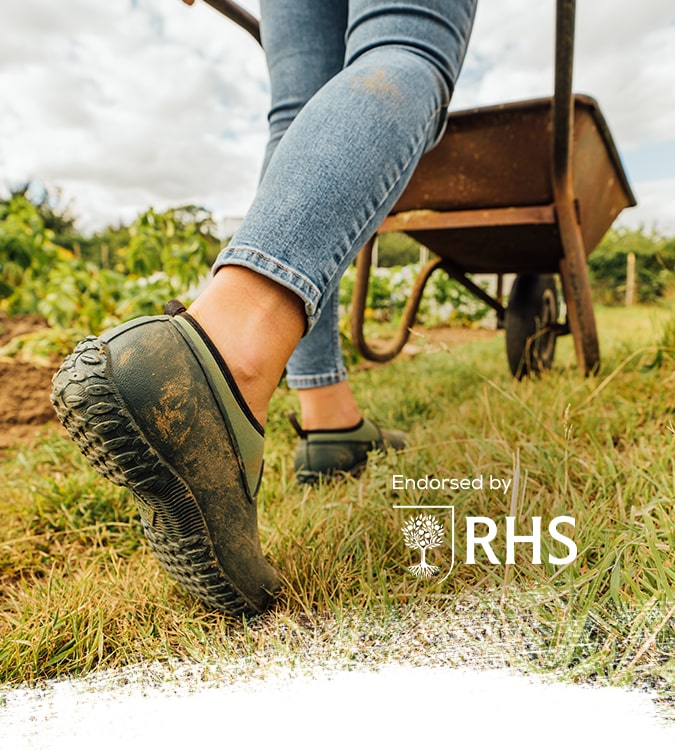 Person walking with a wheelbarrow on grass, wearing a pair of Muck Boots Muckster II Shoes with an RHS logo overlayed.