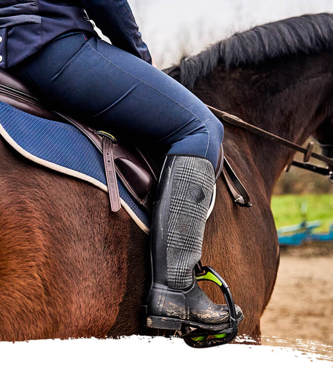 Woman riding a brown horse on a dirt path with a blurred background, she's wearing a pair of Muck Boots Women's Derby Tall Boots