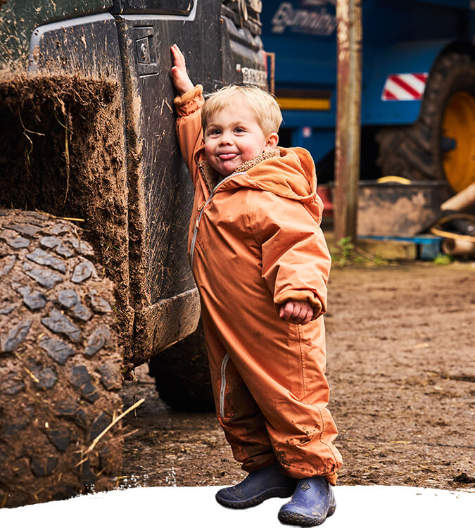Child wearing a pair of blue Muck Boots  Kids Muckster Boots in an orange jumpsuit standing next to a large tire on a dark background