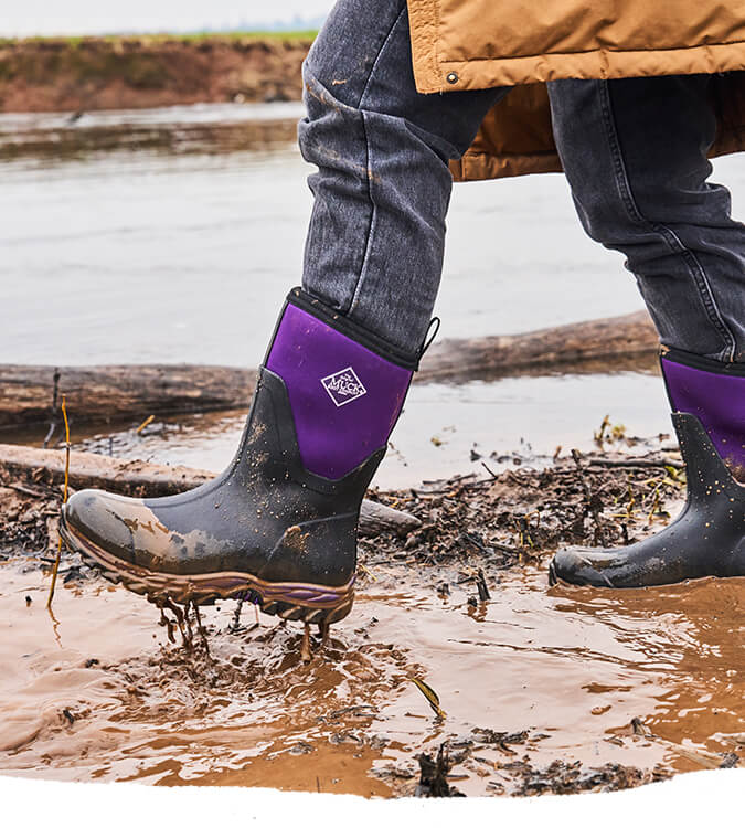 Person wearing a pair of Muck Boots Women's Arctic Sport II boots in black acai, walking through a muddy puddle.
