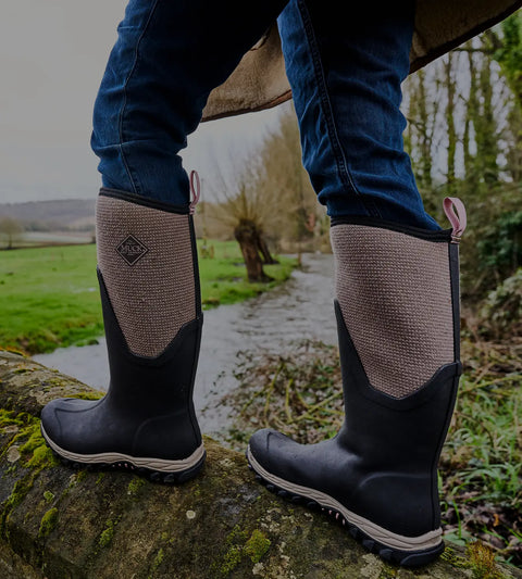 Close-up image of a woman wearing a pair of Muck Boots Women's Arctic Sport II Tall Boots while walking along a moss covered bridge wall.