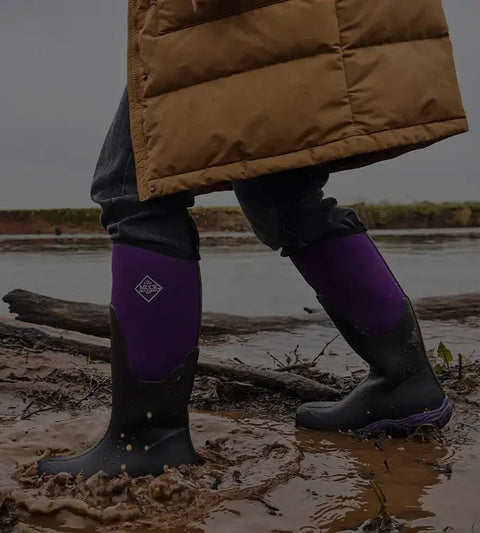 Person walking trough muddy water wearing a pair of Muck Boots Arctic Sport Wellingtons in Black Acai