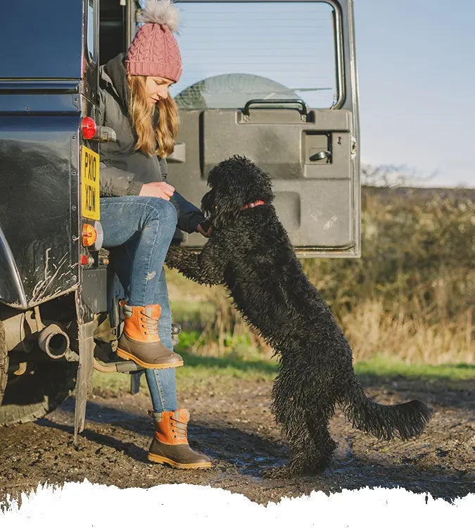 A woman wearing a pair of Tan Muck Boots Muck Originals Duck Lace-Up Leather Short Boots, sat in the back of a Land Rover feeding a black dog.