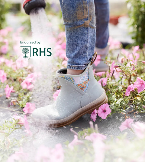 Close-up image of a woman wearing a pair of Muck Boots Muck Originals Pull-On Ankle Boots while watering pink flowers.