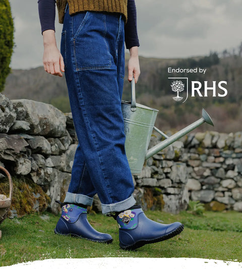 Woman holding a metal watering can wearing a pair of Muck Boots Muckster II Ankle Boots walking in front of a dry stone wall.