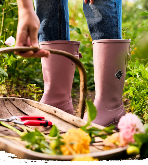 Person wearing a pair of Woodrose twilight mauve Muck Boots Rainscape Tall Boots with a basket of cut flowers in a garden setting