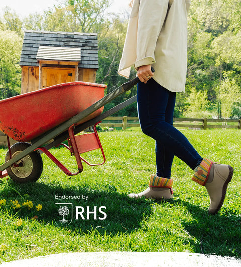 Person wearing a pair of Muck Boots Women's RHS Muckster II Short Boots pushing a red wheelbarrow with a small wooden shed in the background, featuring the RHS logo