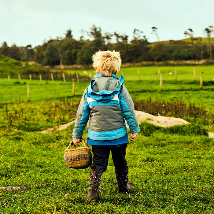 Child wearing a pair of Muck Boots in a blue and grey jacket walking through a grassy field holding a basket.