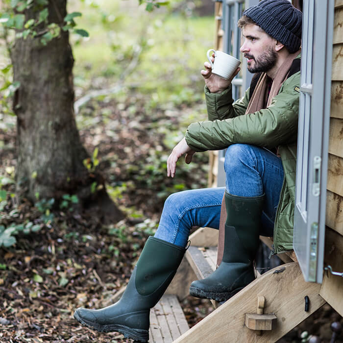 Man sitting on a wooden step outdoors, wearing a pair of green Muck Boots Arctic Outpost wellingtons and a green jacket, holding a white mug.
