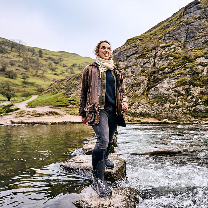 Woman wearing a pair of Muck Boots wellingtons, standing on a rock in a river with mountains in the background.