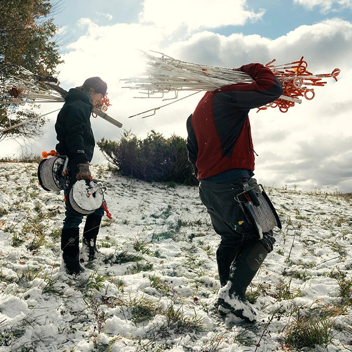 Man and woman carrying rolls of wire and plastic poles. Both are wearing Muck Boots wellingtons in the snow