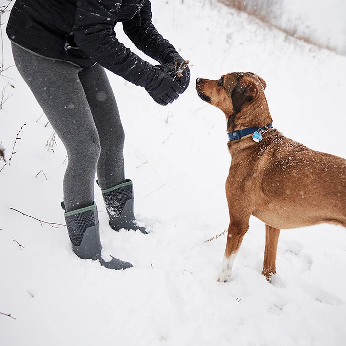 A woman wearing a pair of Muck Boots wellingtons and gloved hands, holding out a treat to a dog in the snow.