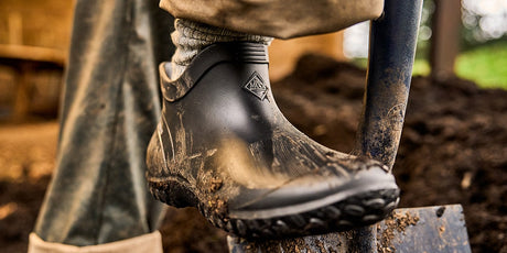 Muddy work boot on a metal step with a blurred outdoor background featuring a pair of Women's Muckster Lite boots.