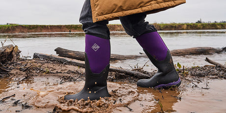 Person wearing a pair of Black Acai Muck Boots Arctic Sport Tall Boots in a muddy outdoor setting with water and logs.