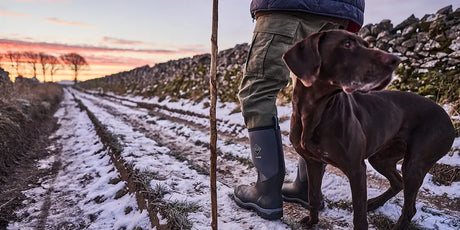 A man wearing a pair of Muck Boots wellingtons stood on a snow covered lane with a dog at sunrise.