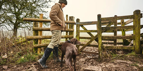 A woman wearing a beanie hat, fleece and pair of Muck Boots Arctic Sport II wellingtons approaching a wooden gate over muddy ground. 