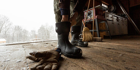 Man putting a pair of Muck Boots Arctic Sport Wellingtons inside a wooden shed with a snowy scene behind