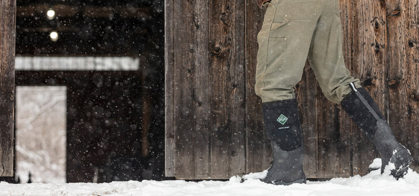 Close-up image of a person wearing a pair of Muck Boots Arctic Sport Wellingtons in the snow outside of a n open wooden barn door