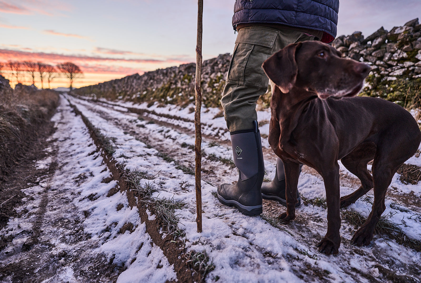 Man with a stick stood on a snow covered country lane, wearing a pair of Muck Boot Calder wellingtons with a dog behind him looking backwards