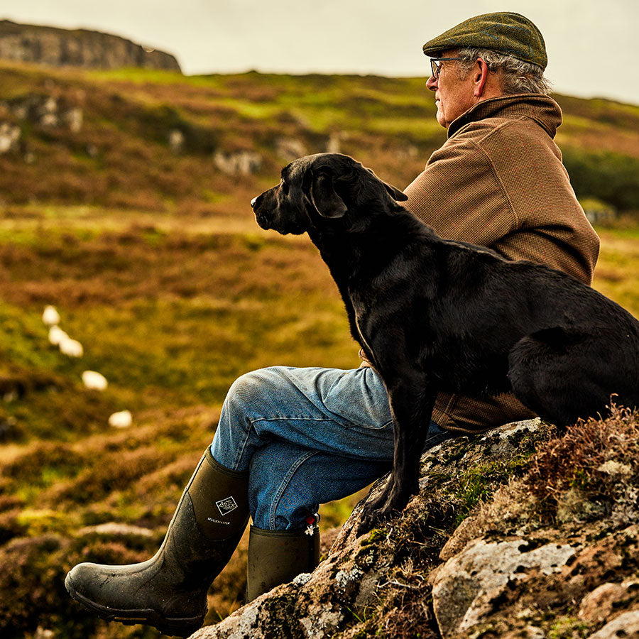 A man wearing a flat cap and pair of Muck Boot wellington boots, sat on a rocky outcrop with a black Labrador, looking across a moorland valley 