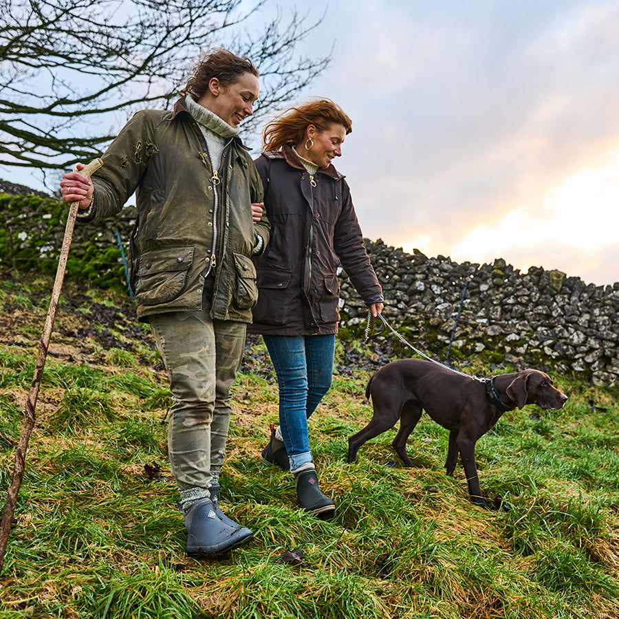 Two women walking a dog in a field, wearing Muck Boots Original ankle boots, with a dry stone wall behind them