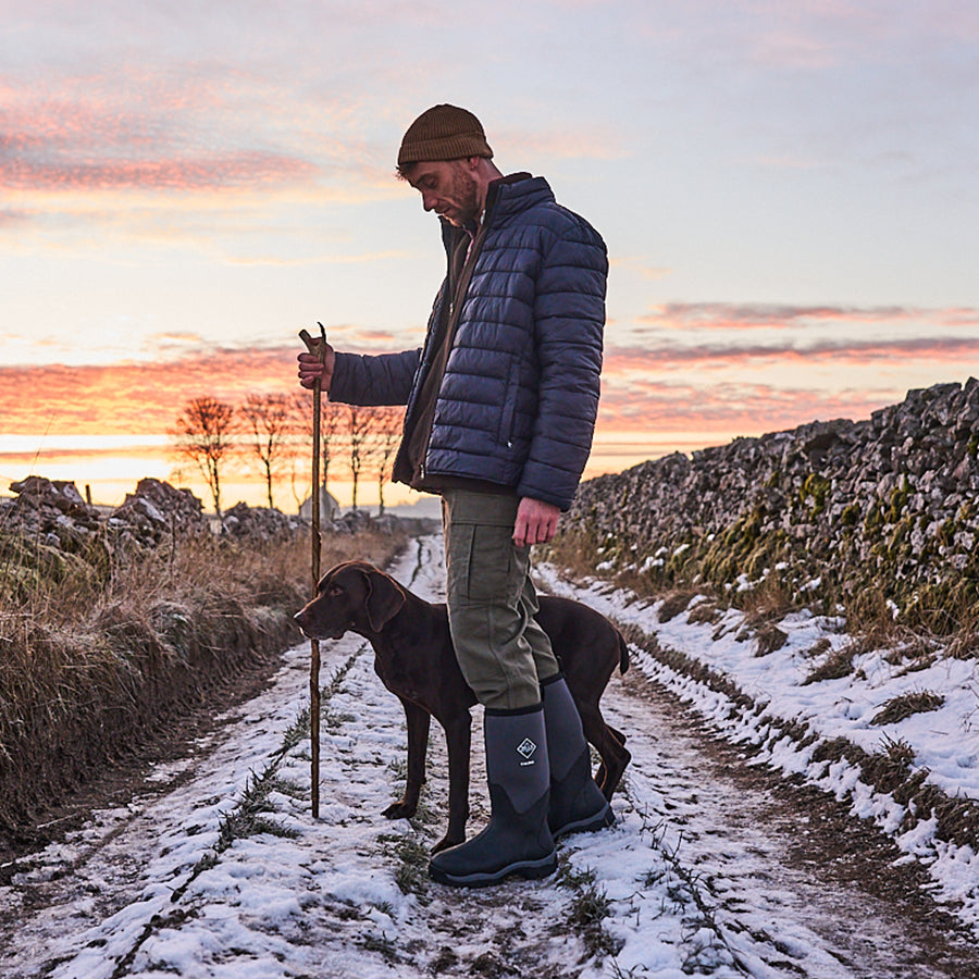 Man wearing a beanie, blue puffer jacket, olive trousers and a pair of Muck Boots. He's holding a stick, with a dog stood next to him on a snow-covered lane at sunrise