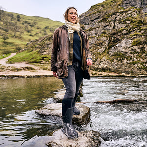 Woman stood on rocks in a river within a valley, wearing a scarf, wax jacket and a pair of Muck Boots wellingtons
