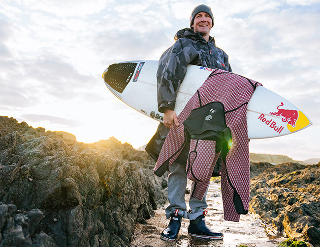 Andrew 'Cotty' Cotton stood on a rocky beach, holding a surfboard with dry suit draped over it. He's wearing a beanie hat, long coat and Xtratuf ankle deck boots