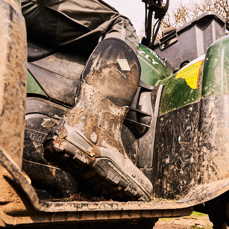 Close-up image of a person wearing a pair of muddy Muck Boots Arctic Sport Wellingtons while sat on a quad bike