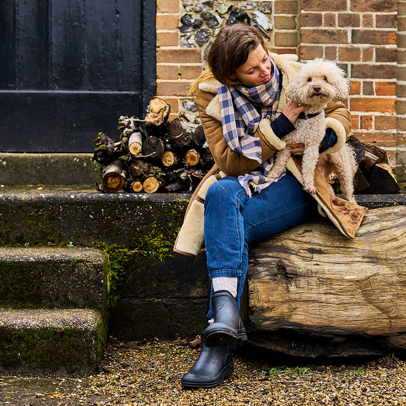 Woman sat on a large log, wearing a pair of black Muck Boots Muck Originals while holding a small dog